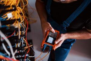 Top view of young man in uniform with measuring device that works with internet equipment and wires in server room photo