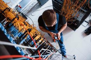 Top view of young man in uniform with measuring device that works with internet equipment and wires in server room photo