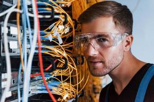 Young man in protective glasses works with internet equipment and wires in server room photo
