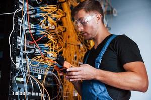 joven uniformado con dispositivo de medición trabaja con equipos de internet y cables en la sala de servidores foto