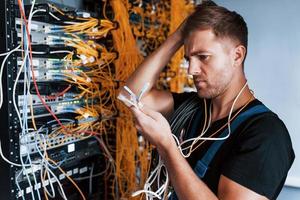 Young man in uniform feels confused and looking for a solution with internet equipment and wires in server room photo