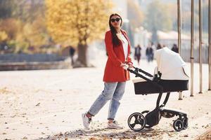 Mother in red coat have a walk with her kid in the pram in the park at autumn time photo