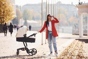 Mother in red coat have a walk with her kid in the pram in the park at autumn time photo