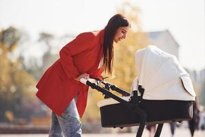 Mother in red coat have a walk with her kid in the pram in the park at autumn time photo