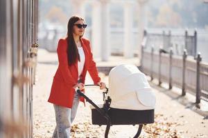 Mother in red coat have a walk with her kid in the pram in the park at autumn time photo