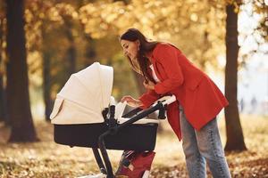 madre con abrigo rojo da un paseo con su hijo en el cochecito en el parque con hermosos árboles en otoño