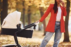 hablando por teléfono. madre con abrigo rojo da un paseo con su hijo en el cochecito del parque en otoño foto