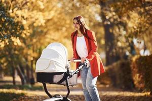 Mother in red coat have a walk with her kid in the pram in the park with beautiful trees at autumn time photo