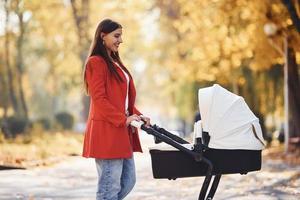 madre con abrigo rojo da un paseo con su hijo en el cochecito en el parque con hermosos árboles en otoño foto