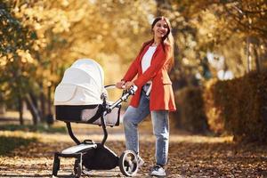 Mother in red coat have a walk with her kid in the pram in the park with beautiful trees at autumn time photo