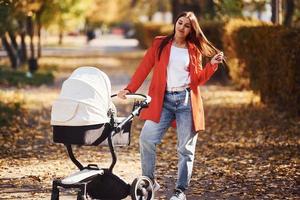 Mother in red coat have a walk with her kid in the pram in the park with beautiful trees at autumn time photo