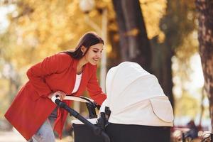 madre con abrigo rojo da un paseo con su hijo en el cochecito del parque en otoño foto