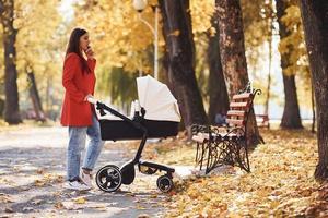 hablando por teléfono. madre con abrigo rojo da un paseo con su hijo en el cochecito del parque en otoño foto