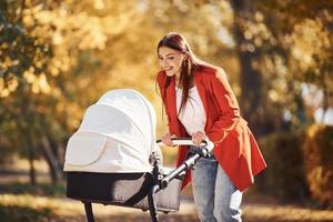 Mother in red coat have a walk with her kid in the pram in the park with beautiful trees at autumn time photo