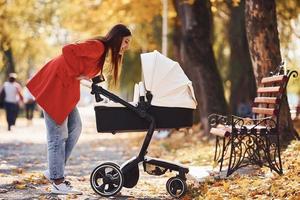 Mother in red coat have a walk with her kid in the pram in the park with beautiful trees at autumn time photo