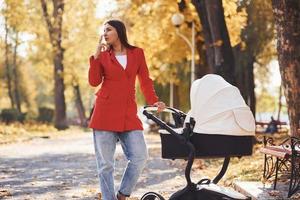 Mother in red coat have a walk with her kid in the pram in the park at autumn time and smoking photo