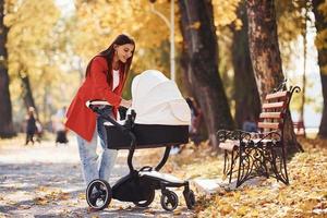 Mother in red coat have a walk with her kid in the pram in the park at autumn time photo