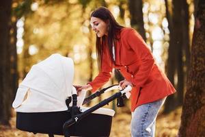 Mother in red coat have a walk with her kid in the pram in the park at autumn time photo