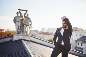 Man in black luxury suit stands on the rooftop against statue photo