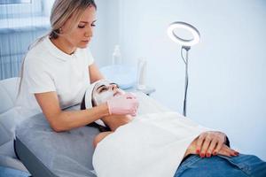 Woman lying down in spa salon and have face cleaning procedure by professional cosmetologist photo