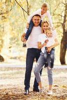 Mother and dad holds kids at shoulders and in hands. Cheerful young family have a walk in an autumn park together photo