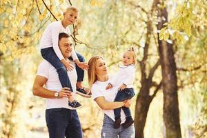 Mother and dad holds kids at shoulders and in hands. Cheerful young family have a walk in an autumn park together photo