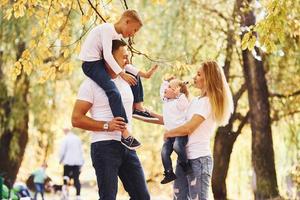 Mother and dad holds kids at shoulders and in hands. Cheerful young family have a walk in an autumn park together photo