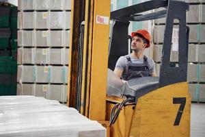 joven trabajador en uniforme se sienta en la carretilla elevadora en el almacén foto