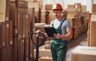 Young male worker in uniform is in the warehouse with notepad and pallet truck photo