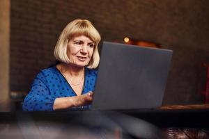 Portrait of senior woman that sits indoors in the cafe with modern laptop photo