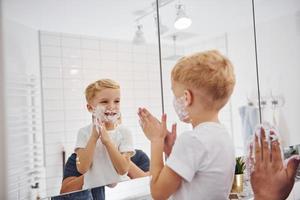 Father with his son is in the bathroom have fun by using shaving gel and looking in the mirror photo