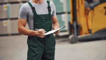 Storage worker in green colored uniform and notepad in hands checks production photo