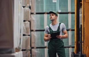 Storage worker in green colored uniform and notepad in hands checks production photo