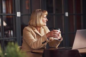 Portrait of senior woman that sits indoors in the cafe with modern laptop and cup of drink in hands photo