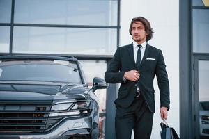 Portrait of handsome young businessman in black suit and tie outdoors near modern car and with shopping bags photo