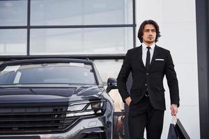 Portrait of handsome young businessman in black suit and tie outdoors near modern car and with shopping bags photo