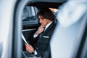 Using notepad. Young businessman in black suit and tie inside modern automobile photo