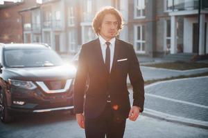 Portrait of handsome young businessman in black suit and tie outdoors near modern car in the city photo