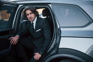 Walks out from car. Young businessman in black suit and tie inside modern automobile photo