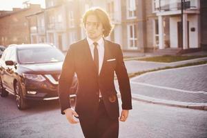 Portrait of handsome young businessman in black suit and tie outdoors near modern car in the city photo