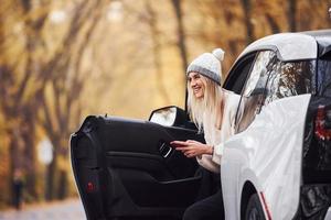 usando un teléfono inteligente. chica tiene viaje de otoño en coche. automóvil nuevo y moderno en el bosque foto