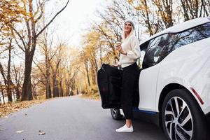 With cup of drink in hands. Girl have autumn trip by car. Modern brand new automobile in the forest photo