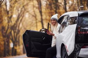 usando un teléfono inteligente. chica tiene viaje de otoño en coche. automóvil nuevo y moderno en el bosque foto