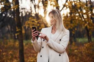 retrato de una joven morena con teléfono en la mano que está en el bosque de otoño durante el día
