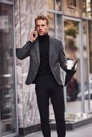 Man in elegant formal wear with cup of drink and notepad in hands is outside against modern building have a conversation by the phone photo