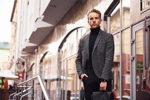 Man in elegant formal wear and with bag is outside against modern building photo