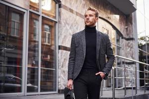 Man in elegant formal wear and with bag is outside against modern building photo