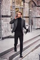 Man in elegant formal wear with cup of drink and notepad in hands is outside against modern building have a conversation by the phone photo