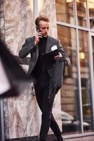 el hombre con ropa formal elegante con una taza de bebida y un bloc de notas en las manos está afuera contra un edificio moderno conversando por teléfono foto