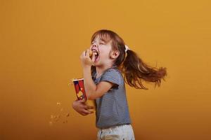 Cute little girl in in the studio eats popcorn and stands against yellow background photo